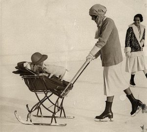 Kinderwagen op ski's (slee) op het ijs, voortgeduwd door moeder op schaatsen. Moeder draagt (onpraktische) schaatskleding en de baby vermaakt zich met een speelgoed varkentje. St. Moritz, Zwitserland, 1926.